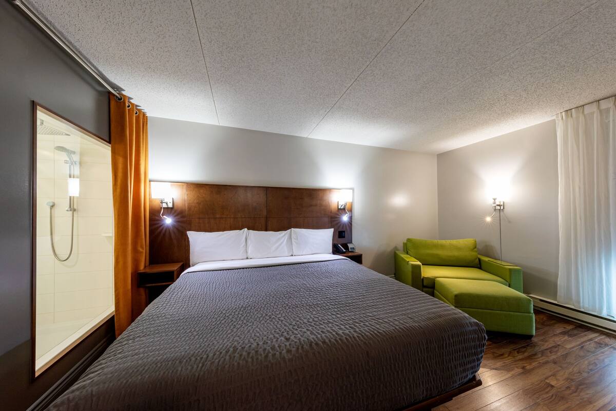 A hotel room with a large bed, a green armchair, a wooden headboard, a nightstand with a lamp, and a view of the outdoors through the window.