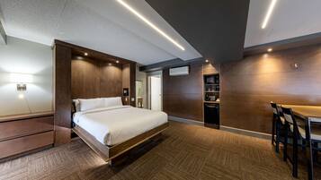 A modern hotel room with a large bed, built-in wardrobe, and a dining area with chairs.