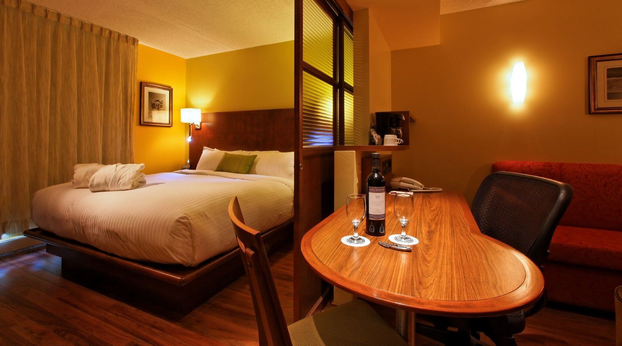 A hotel room with a bed, a desk with a chair, a table, a red sofa, and a lamp.