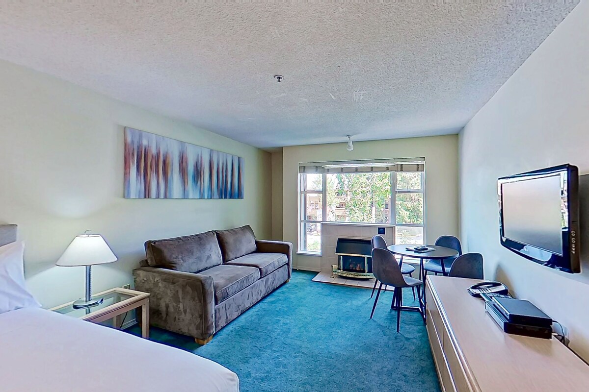 A hotel room with a bed, a sofa, a desk with a television, a fireplace, and a large window.