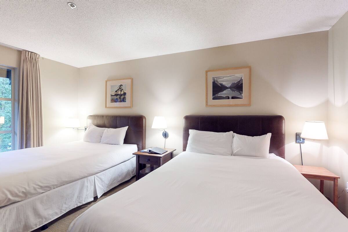 A hotel room with two beds, a nightstand, a lamp, and two framed pictures on the wall.