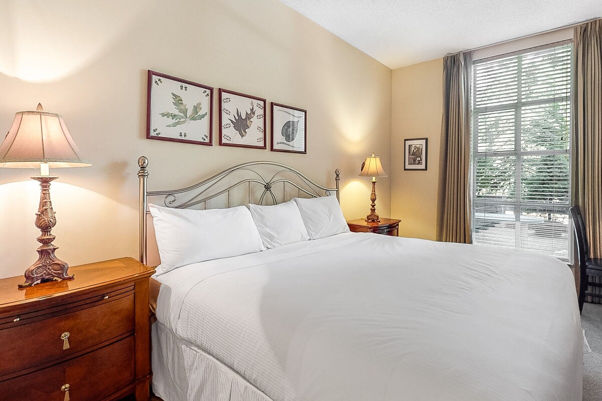 A neatly made bed in a hotel room with a large window, two bedside tables with lamps, and three framed pictures on the wall.