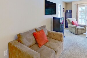 Una sala de estar con un sofá color beige, un cojín rojo, un televisor en la pared, un mueble oscuro y una ventana con cortinas.