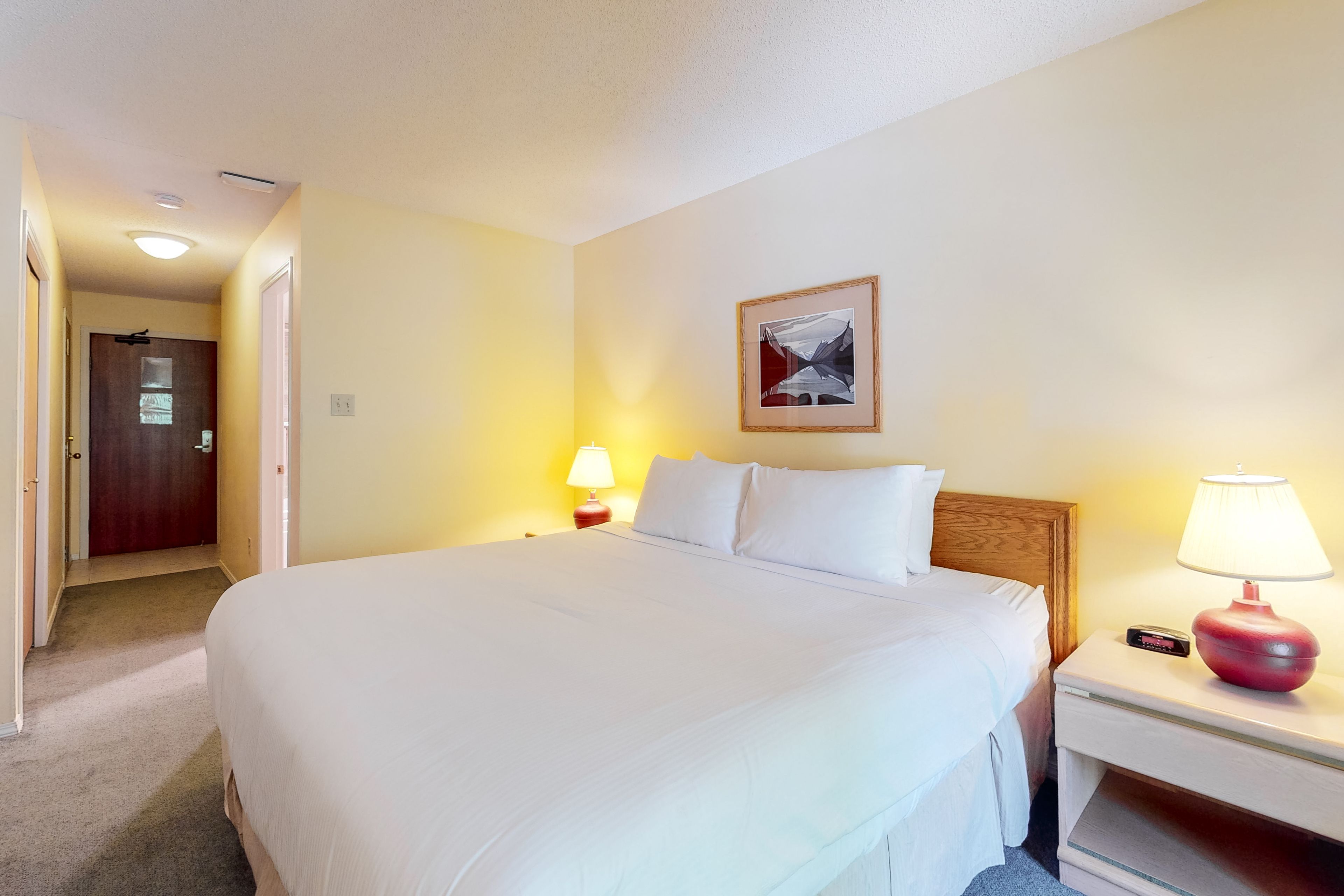 Habitación de hotel con una cama grande, una lámpara de noche, un cuadro en la pared y una puerta que da a otra habitación.