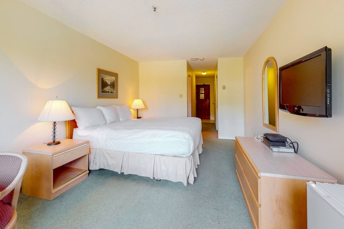 A hotel room with a bed, bedside tables, a desk, a chair, a television, and a door leading to another room.