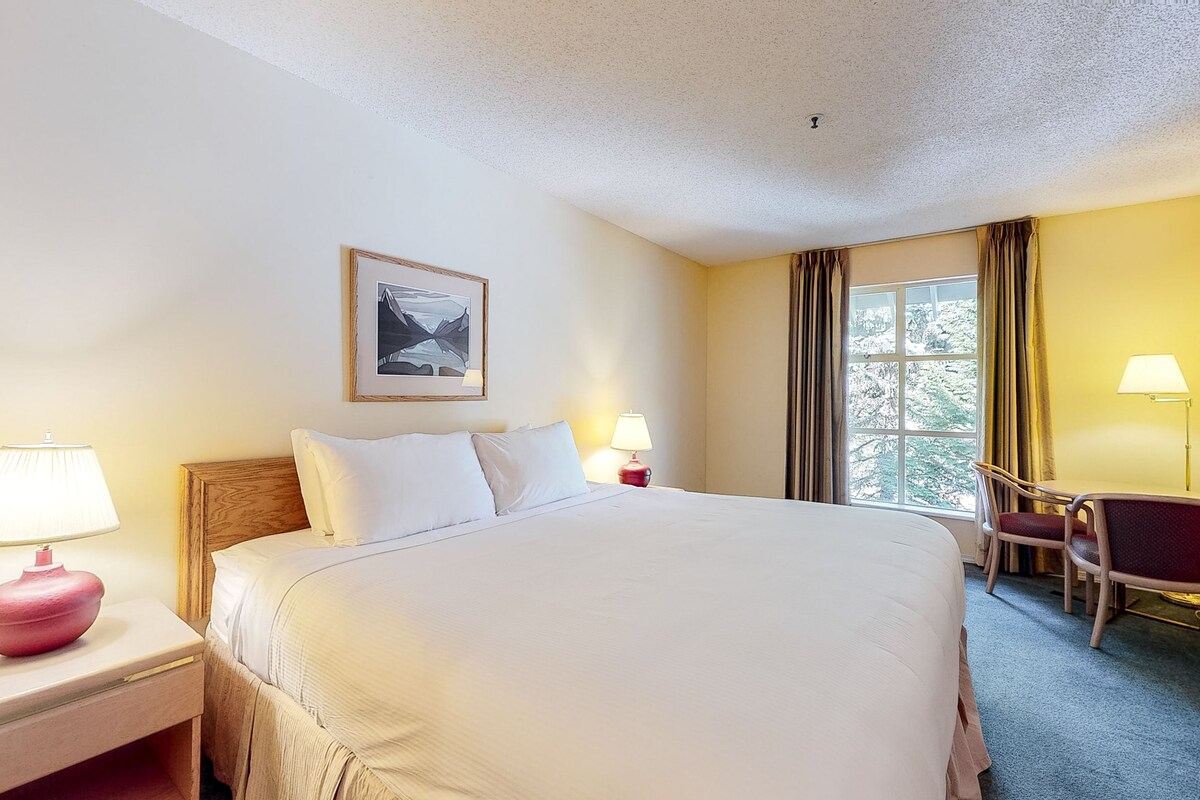 A hotel room with a large bed, two bedside lamps, a chair, a desk, and a window with curtains.