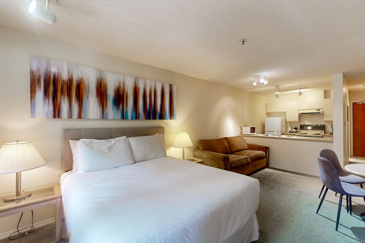A hotel room with a large bed, a desk with a lamp, a chair, a sofa, and a kitchenette in the background.