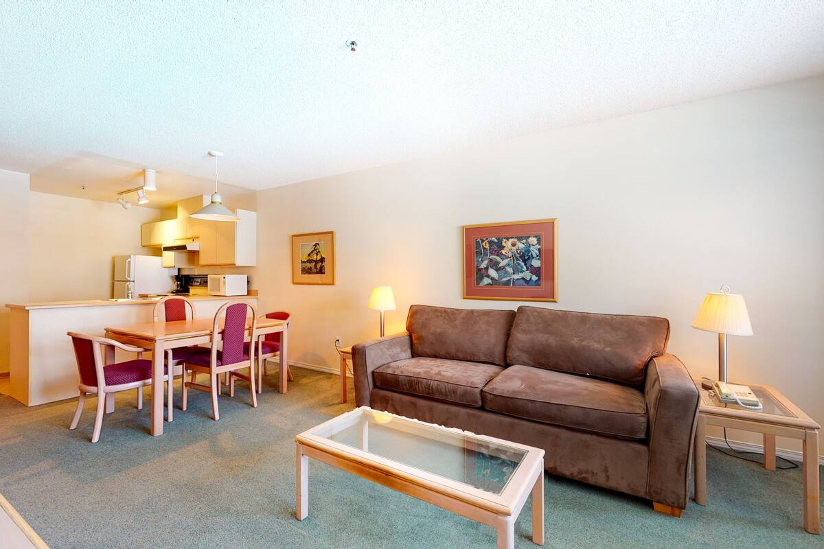 A living room with a brown leather sofa, a glass coffee table, a dining table with chairs, a kitchen area, and two lamps.