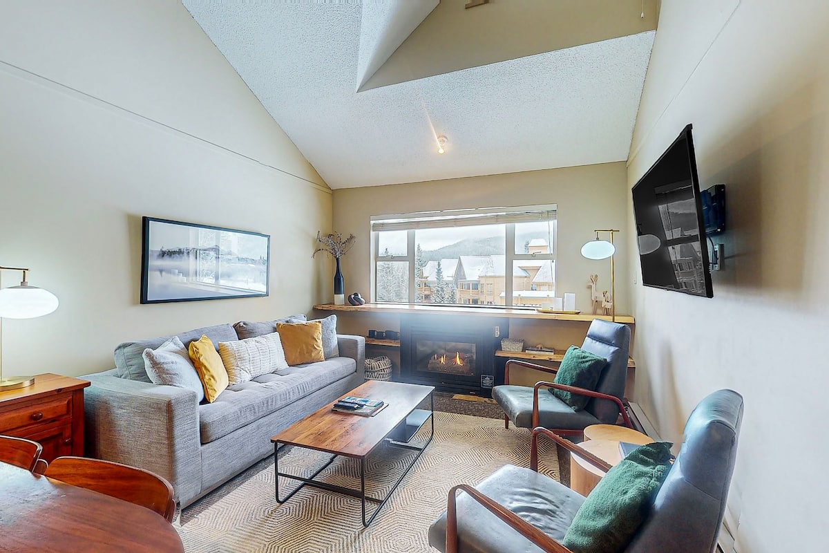 A modern living room with a fireplace, comfortable seating, and a large window offering a view of buildings.