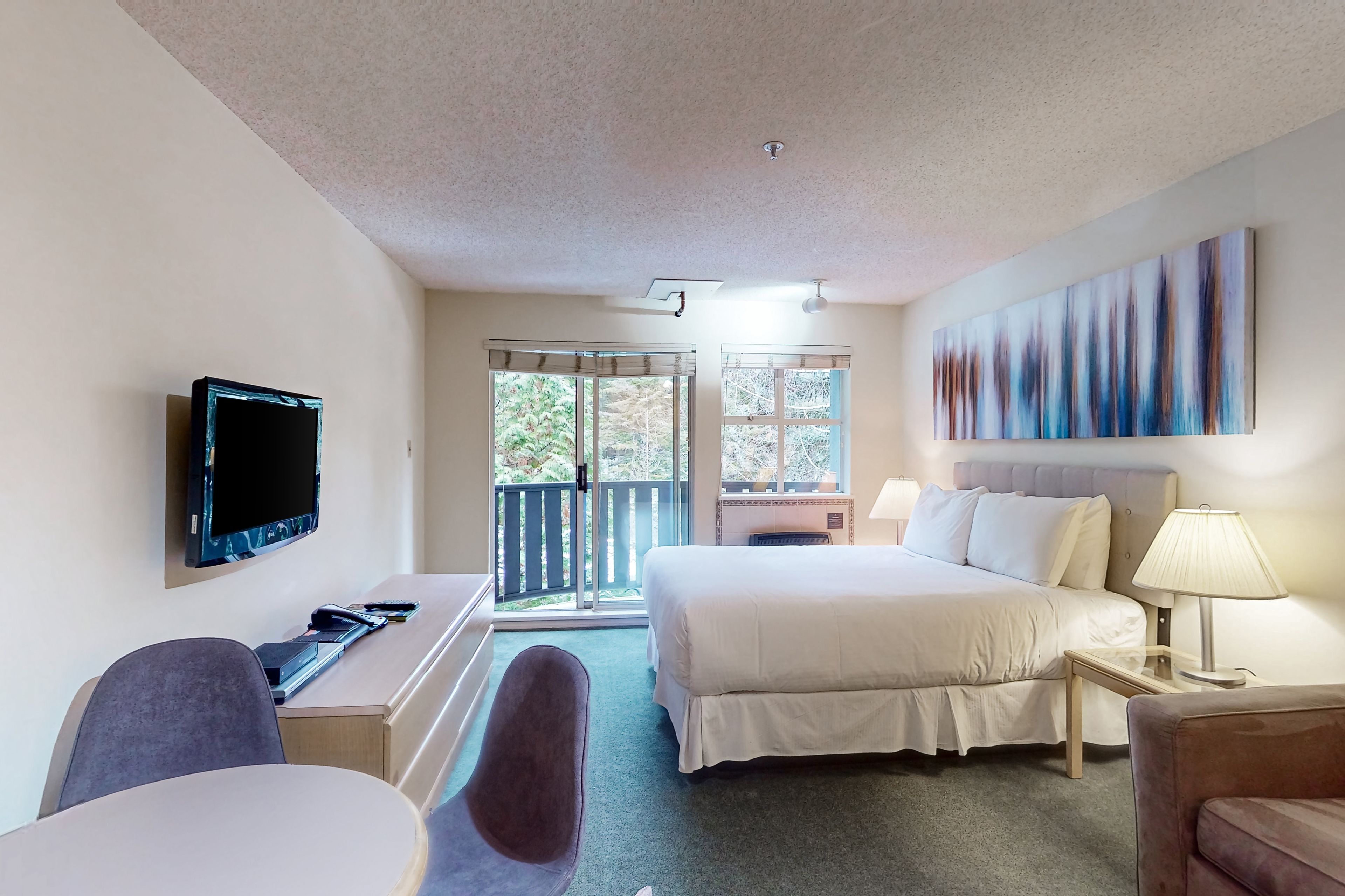A hotel room with a bed, a desk with a computer, a TV, and a window with a view of greenery.