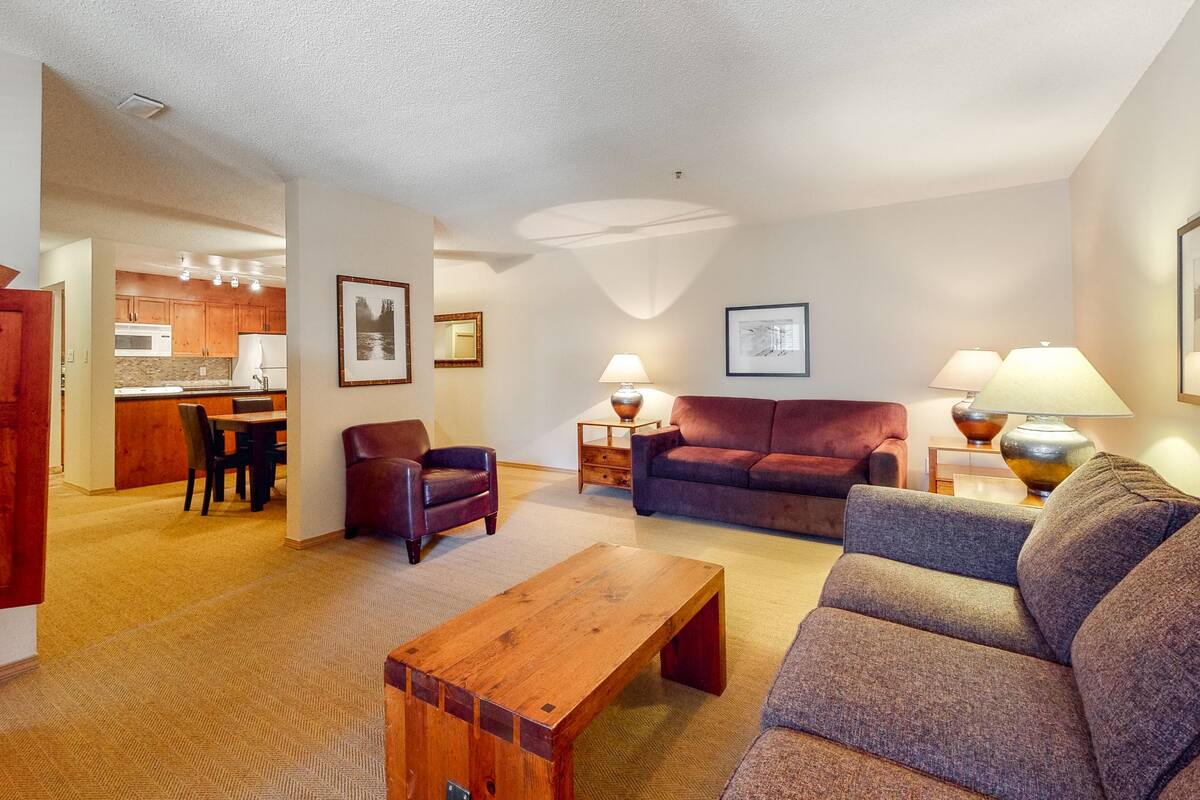 A spacious living room with a wooden coffee table, a grey sofa, and a red armchair. There is a kitchen in the background with wooden cabinets and a dining area with a table and chairs.