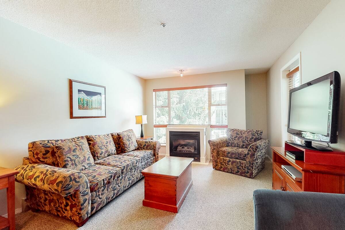A living room with patterned sofas, a wooden coffee table, a fireplace, a television, and a framed picture on the wall.