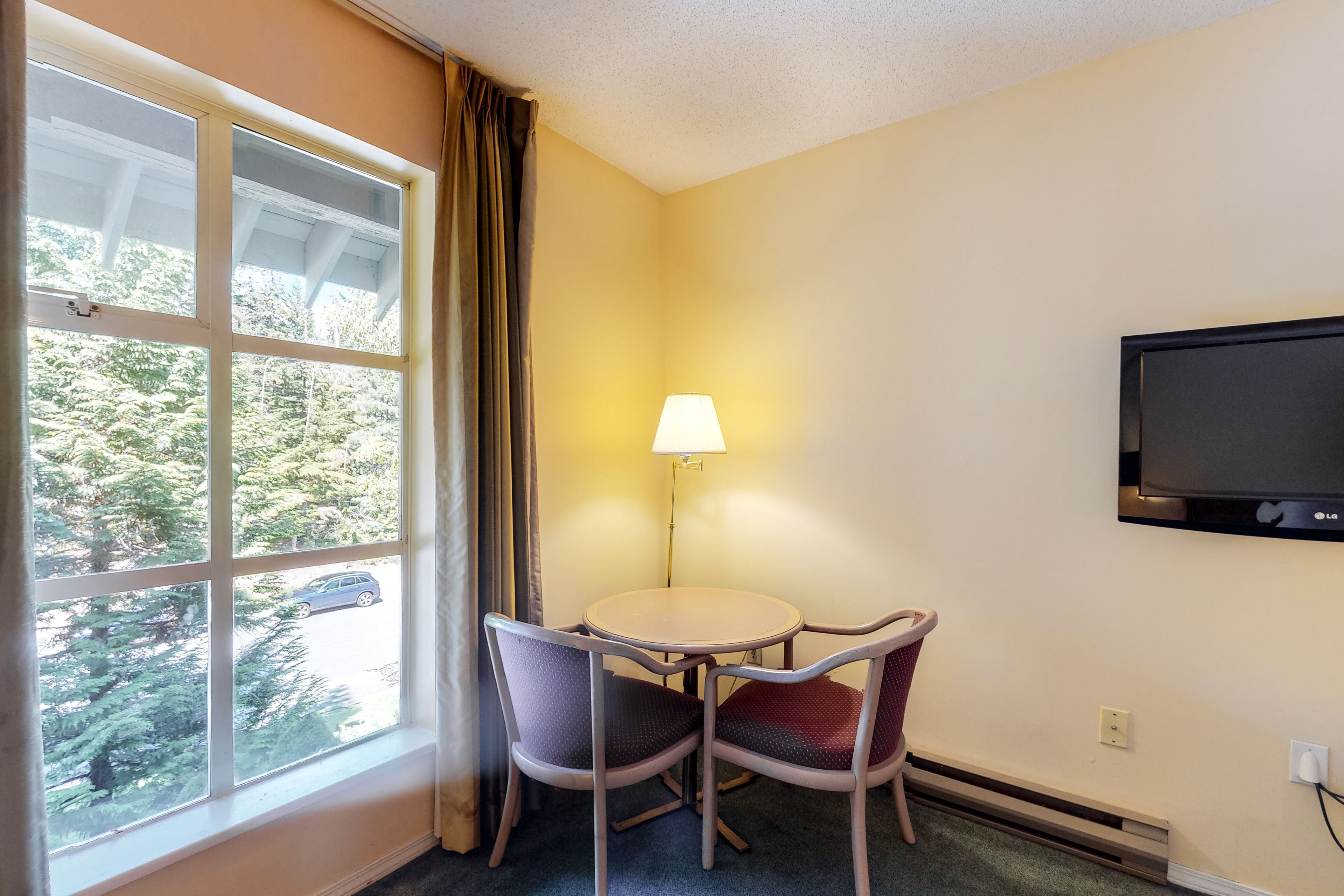 A hotel room with a window, a table with two chairs, a lamp, and a TV mounted on the wall.