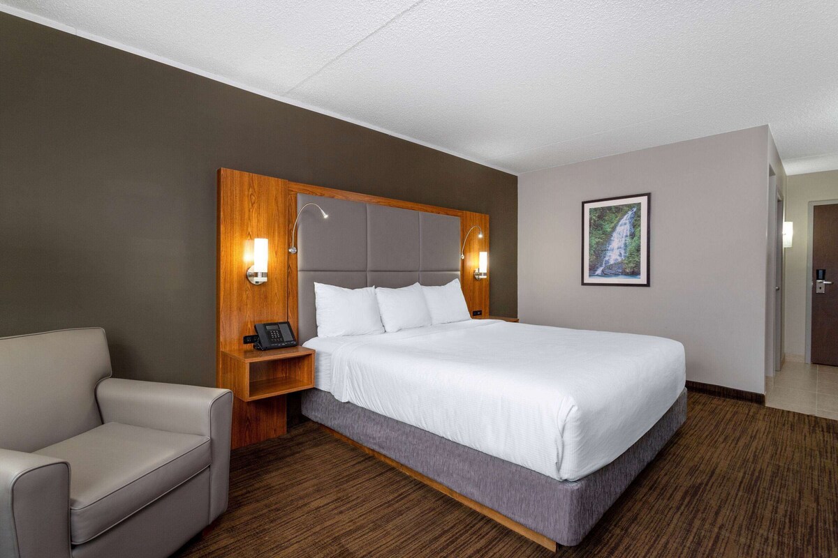 A hotel room with a large bed, a wooden headboard, a bedside table with a lamp, a chair, and a framed picture on the wall.