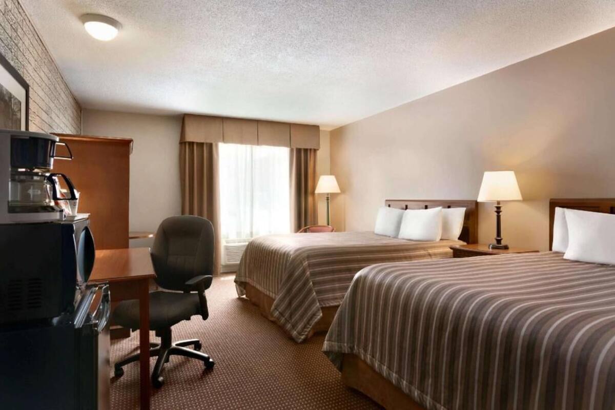 A hotel room with two beds, a desk, a chair, a coffee maker, and a lamp.