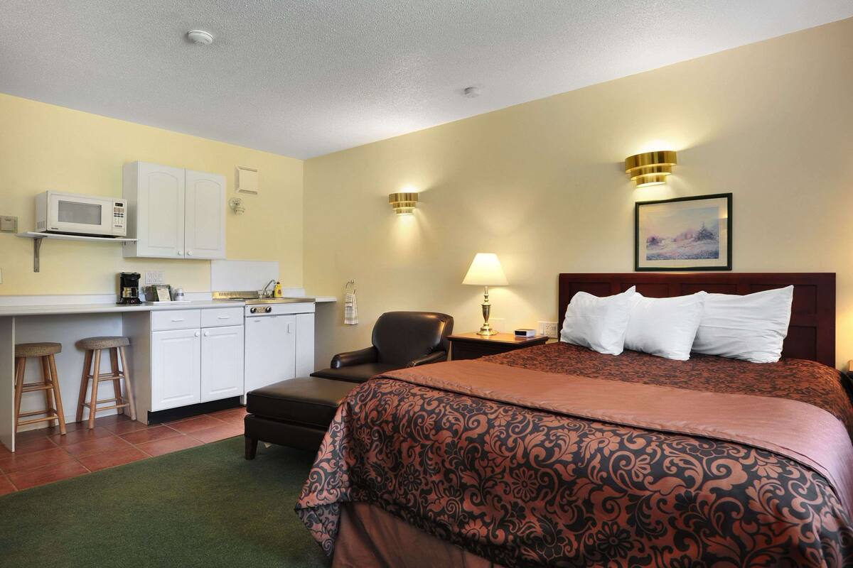A hotel room with a bed, a desk with a microwave, a chair, a lamp, and a picture on the wall.
