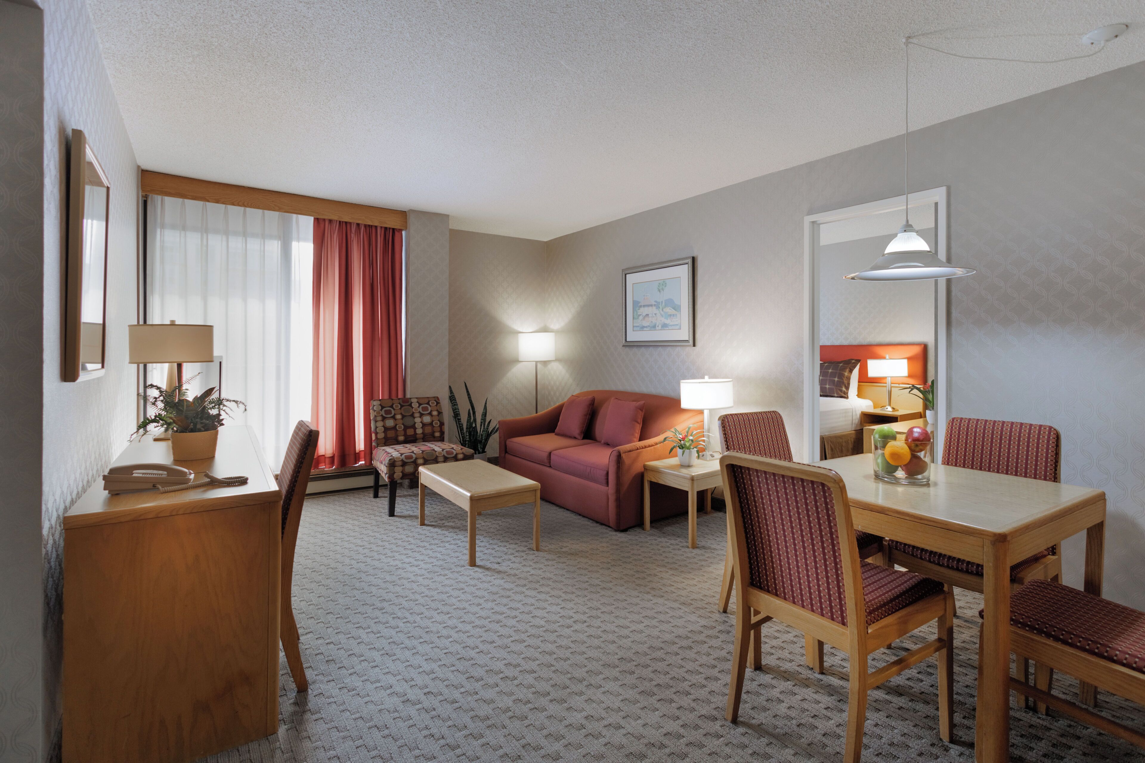 A hotel room with a sofa, a dining table with chairs, a desk with a lamp, and a large window with curtains.