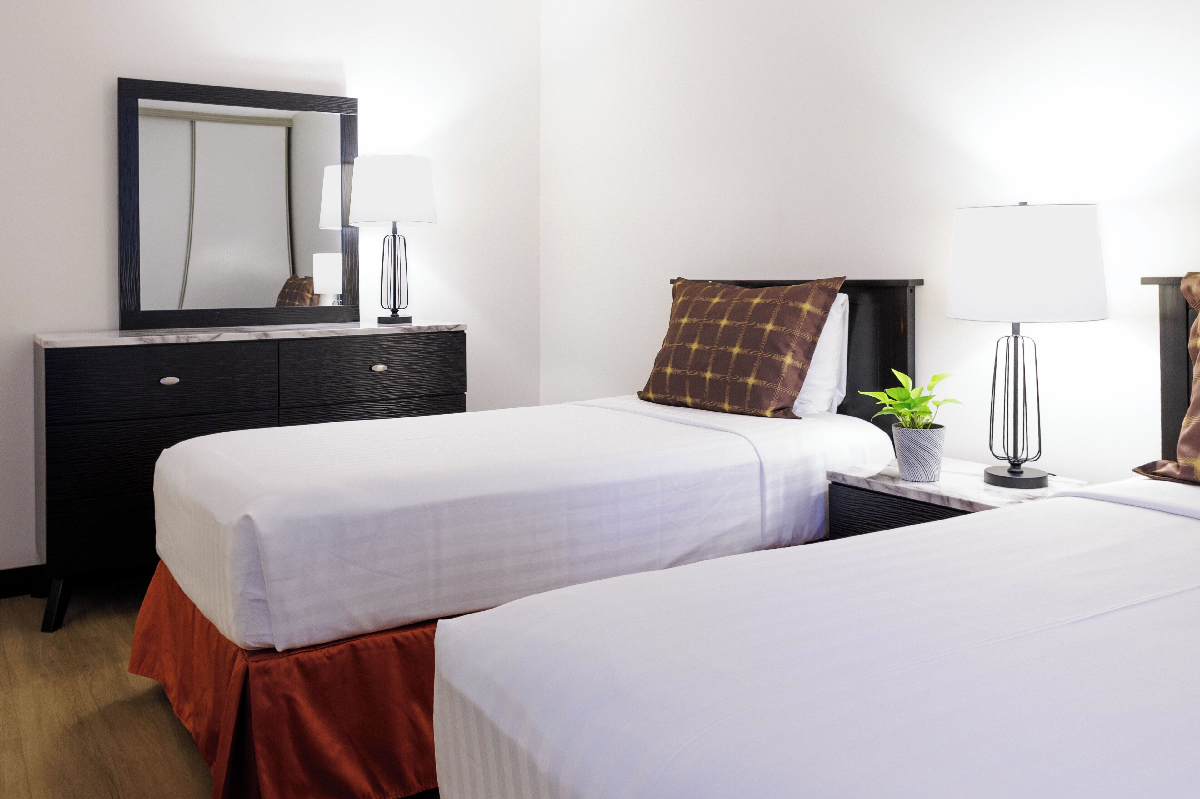 A hotel room with two beds, a bedside table with a lamp, a mirror, and a potted plant.