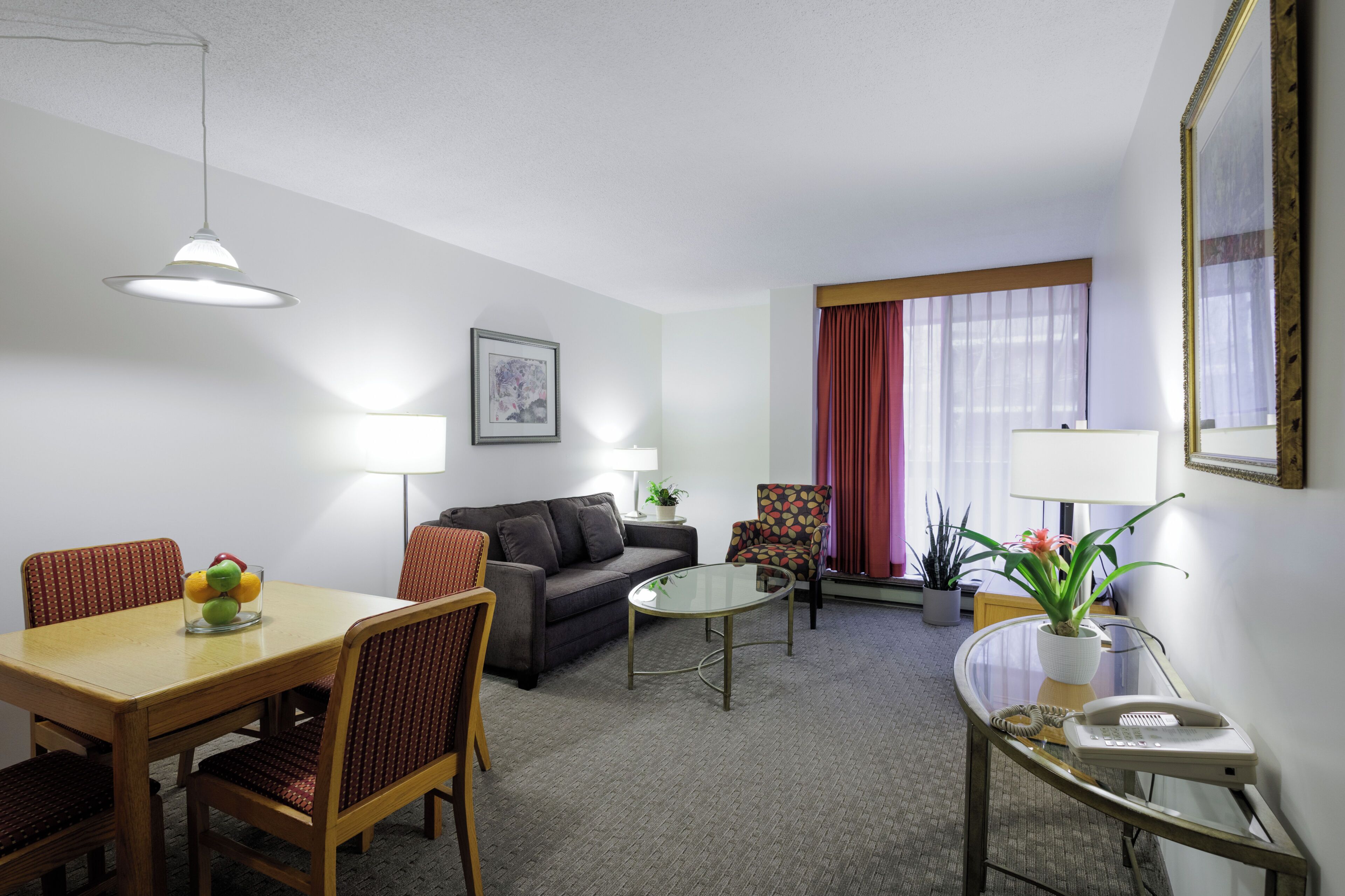A hotel room with a dining table, two couches, a coffee table, a chair, a lamp, a potted plant, and a telephone.