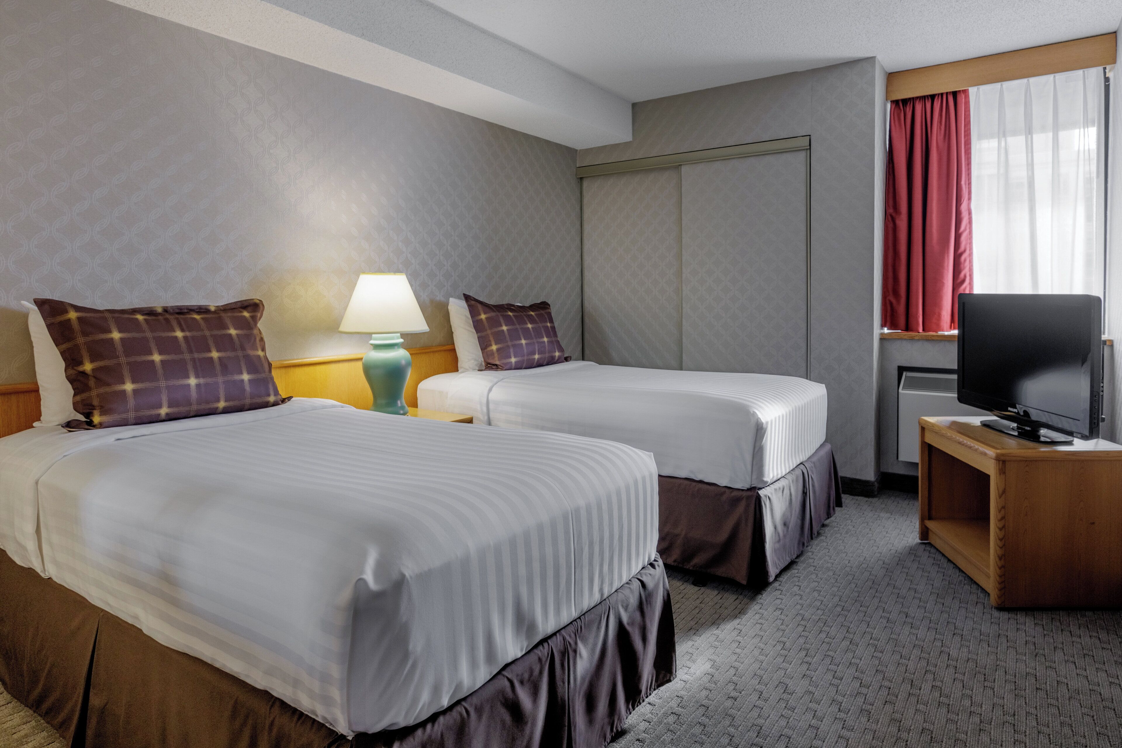 A hotel room with two beds, a television, a lamp, and a window with curtains.