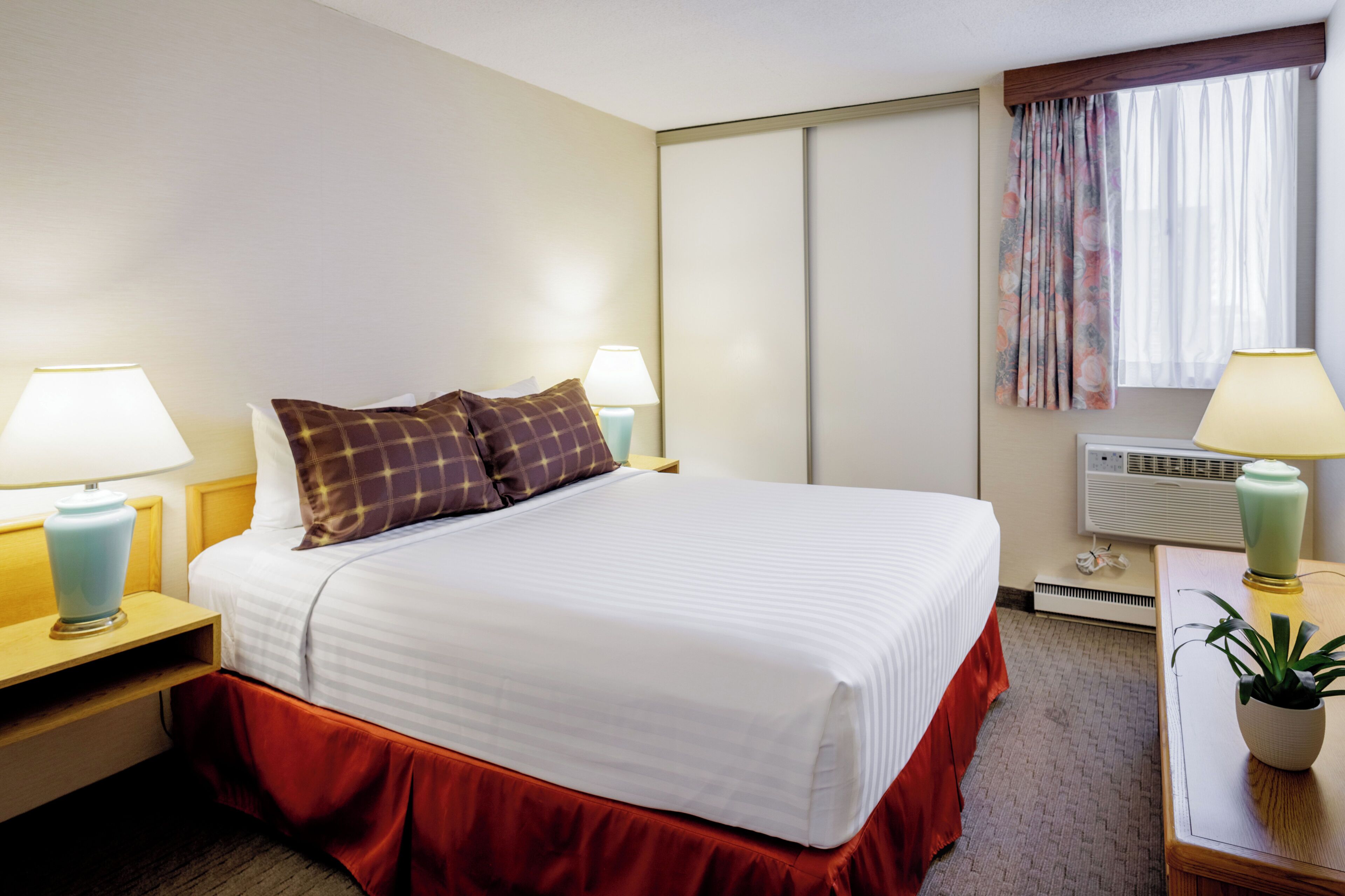 A hotel room with a bed, two bedside lamps, a nightstand, a desk with a lamp, a potted plant, and a window with curtains.