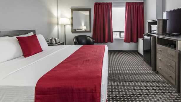 A hotel room with a bed, a desk with a television, a chair, and a window with curtains. - Paradise Inn & Conference Centre (Grande Prairie)