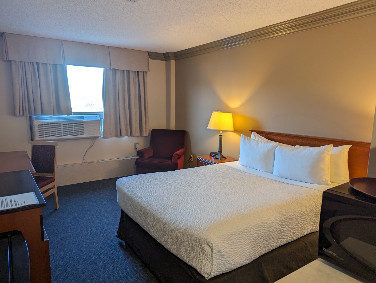 A hotel room with a large bed, a desk, a chair, an air conditioning unit, a lamp, and a television.