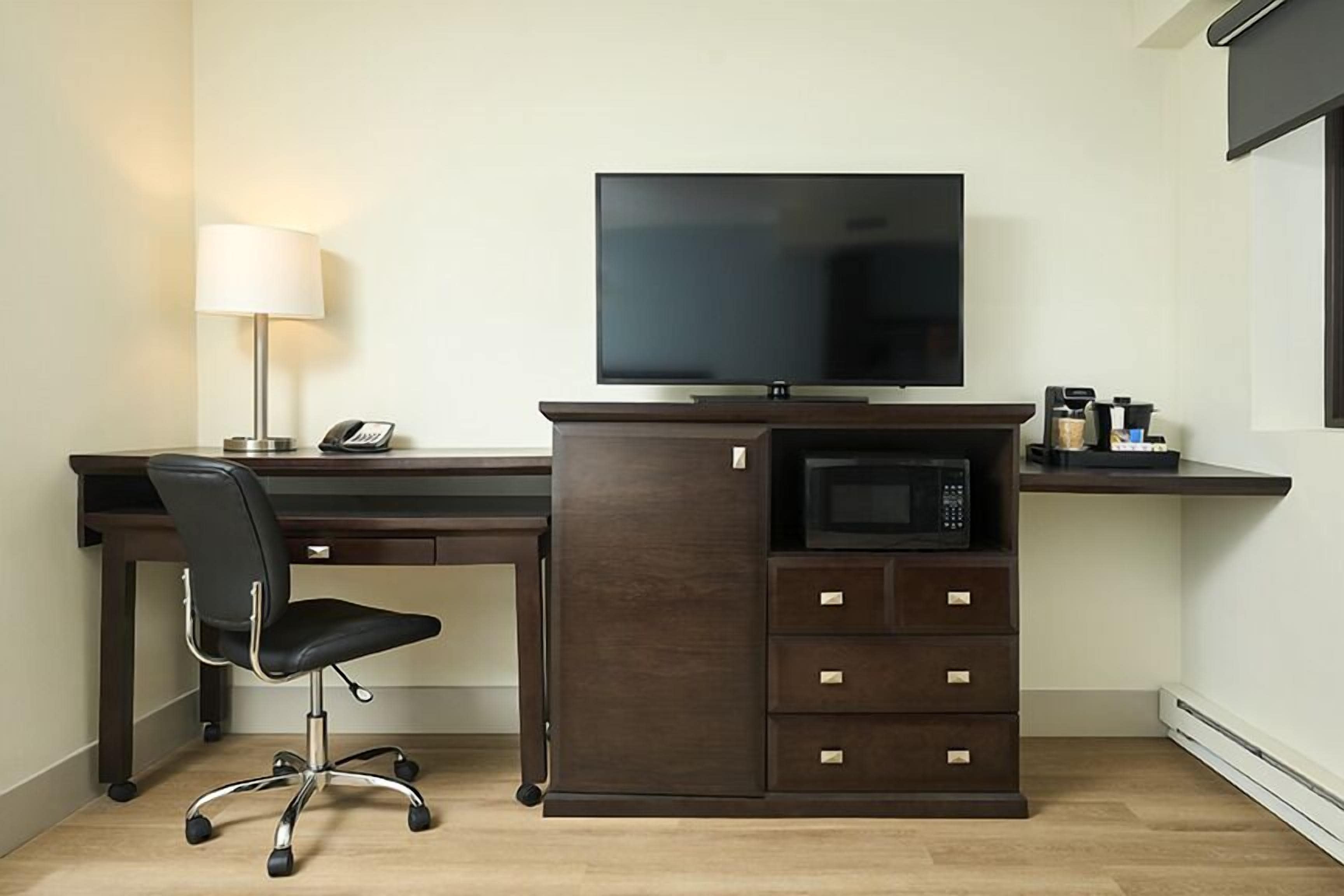 A hotel room with a dark wood desk, a black office chair, a flat-screen TV, a lamp, a microwave, and a coffee maker.