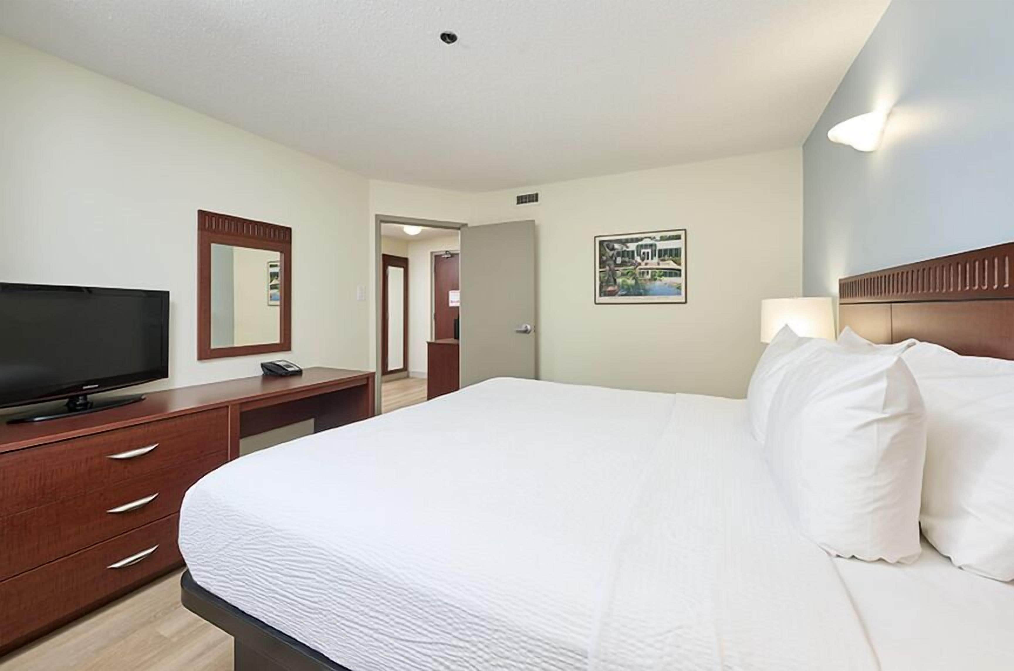 Une chambre d’hôtel avec un grand lit, un téléviseur posé sur une commode en bois, un miroir et un tableau encadré au mur.
