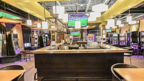 A gaming area with multiple arcade machines, a central counter, and overhead monitors displaying games.