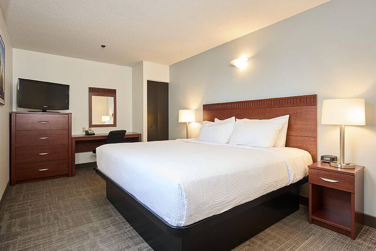 A hotel room with a large bed, a desk with a chair, a TV, a mirror, and two bedside lamps.