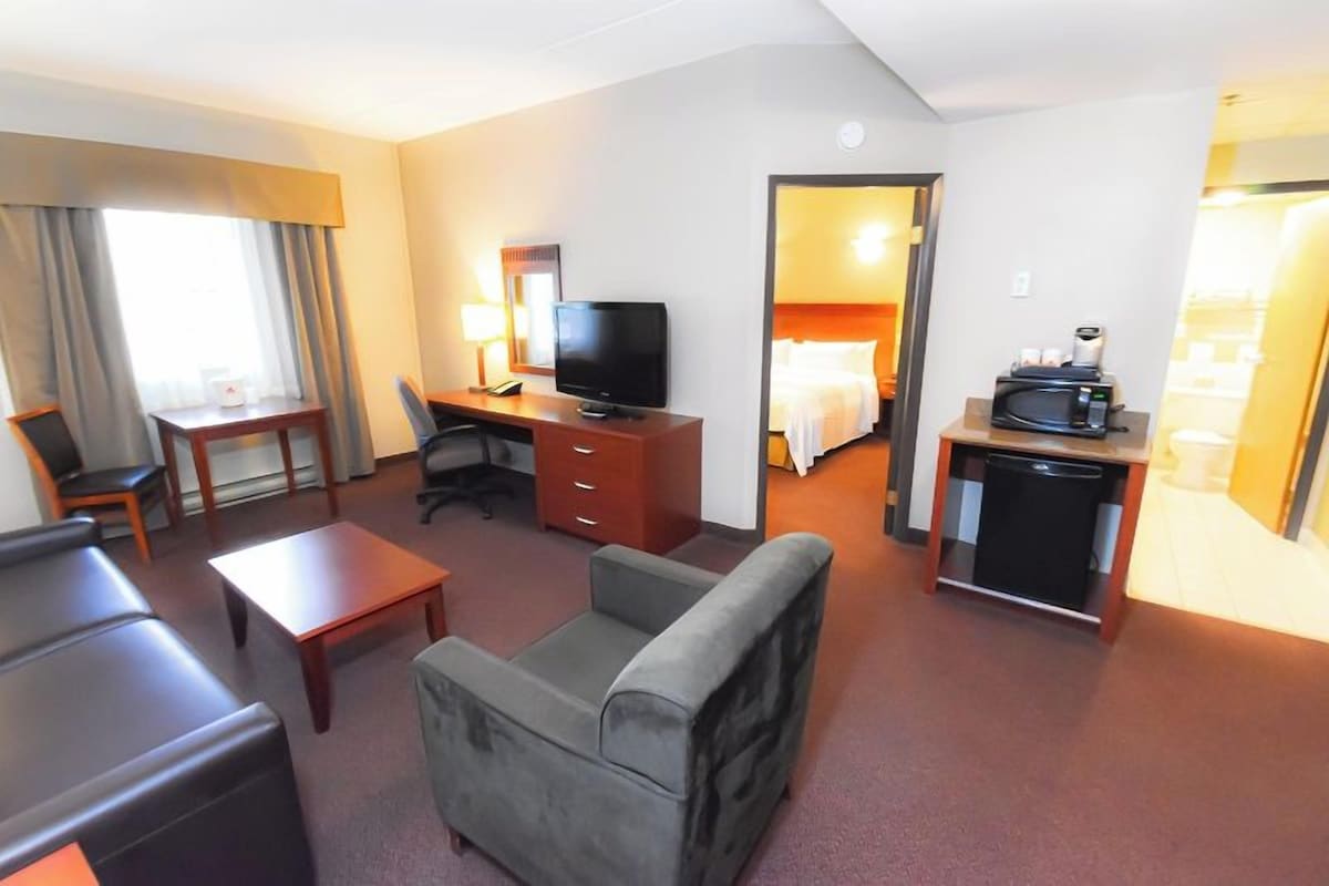 Una habitación de hotel con una cama, un escritorio con televisor, cafetera, una silla, un sofá y una mesita.