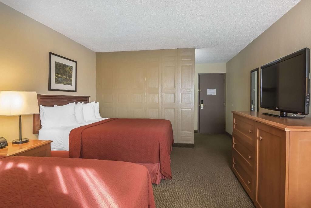 A hotel room with two beds, a TV on a wooden dresser, a lamp on a nightstand, and a door leading to another room.