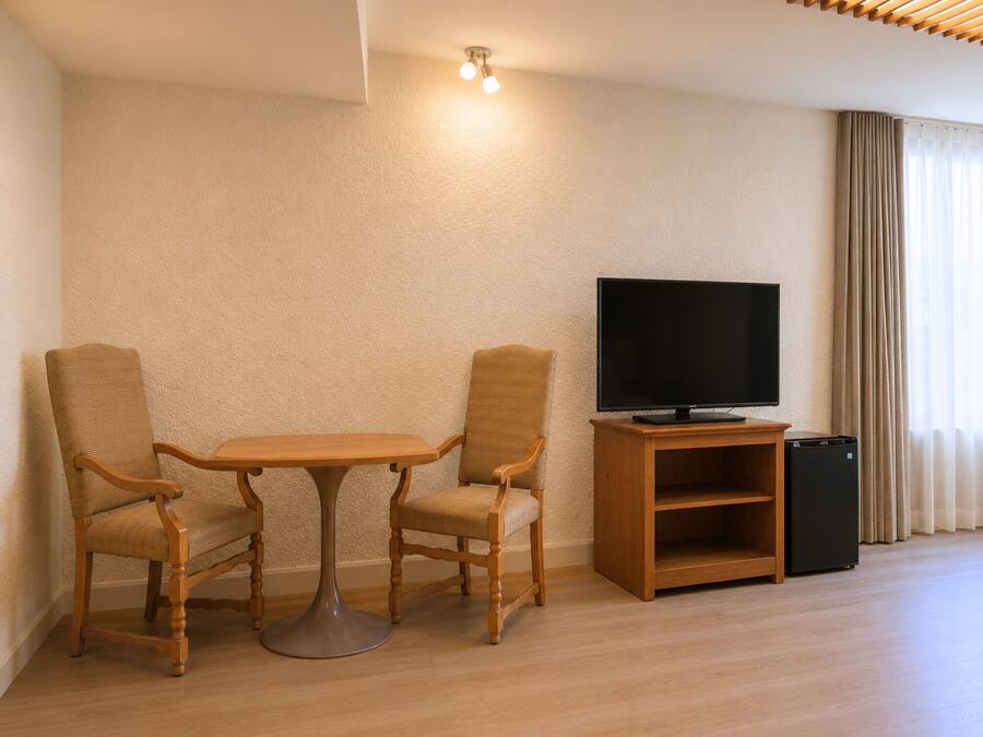 A room with a wooden table and two chairs, a television on a stand, and a trash bin.