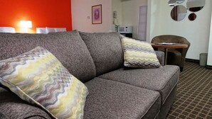 Une chambre d’hôtel moderne avec un canapé gris, des coussins à motifs, une petite table ronde et un four à micro-ondes en arrière-plan.
