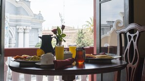 Una mesa de desayuno con platos de comida, una jarra y vasos de jugo de naranja, frente a una vista de un edificio con arquitectura ornamentada.