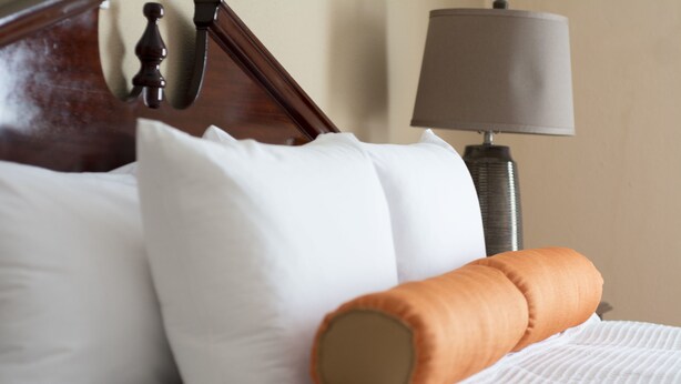 Una cama bien hecha con almohadas blancas y marrones, un cabecero de madera y una lámpara en la mesita de noche.
