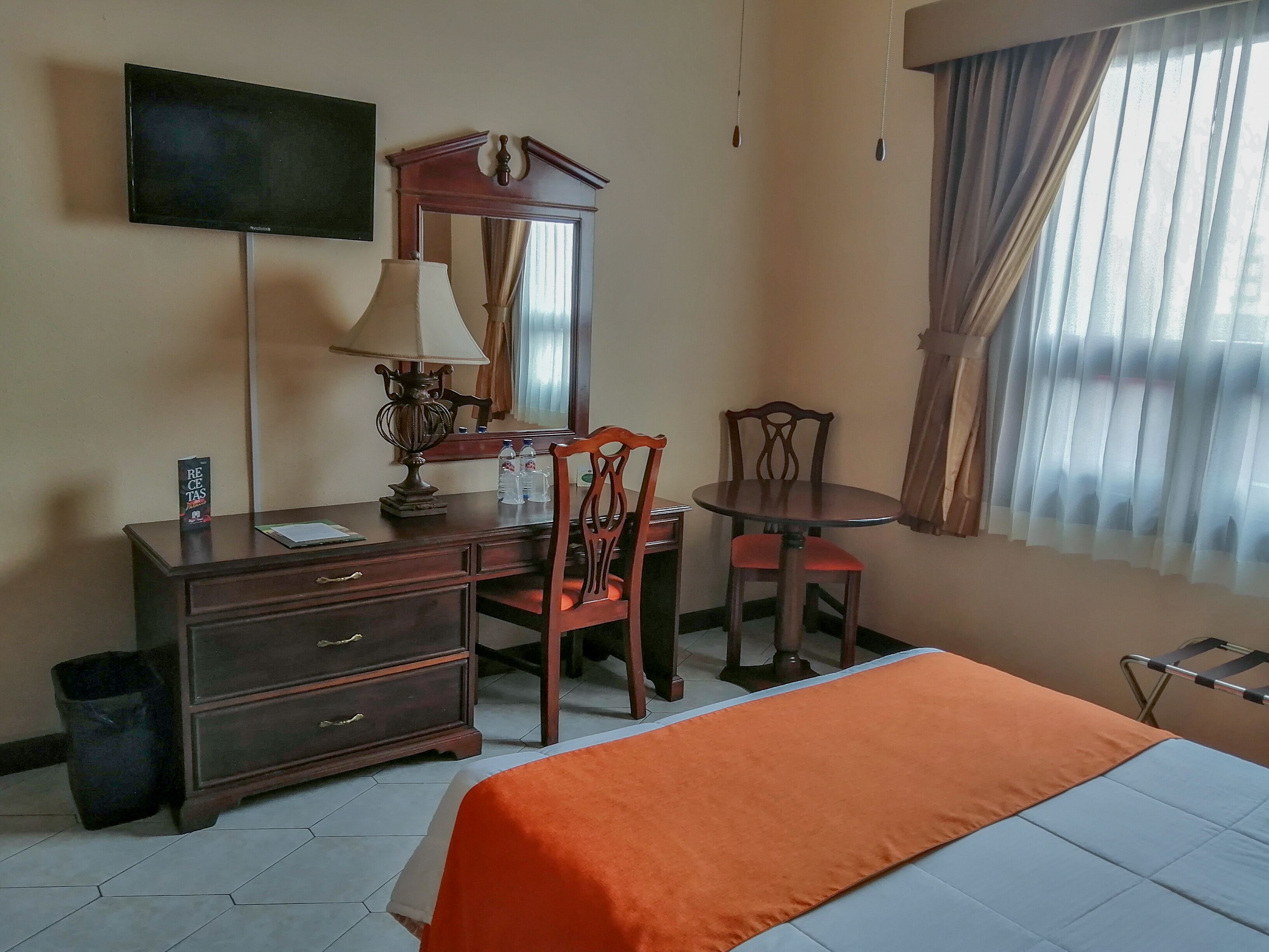 A hotel room with a bed, a desk with a lamp, a chair, a dresser, a television, and a window with curtains.