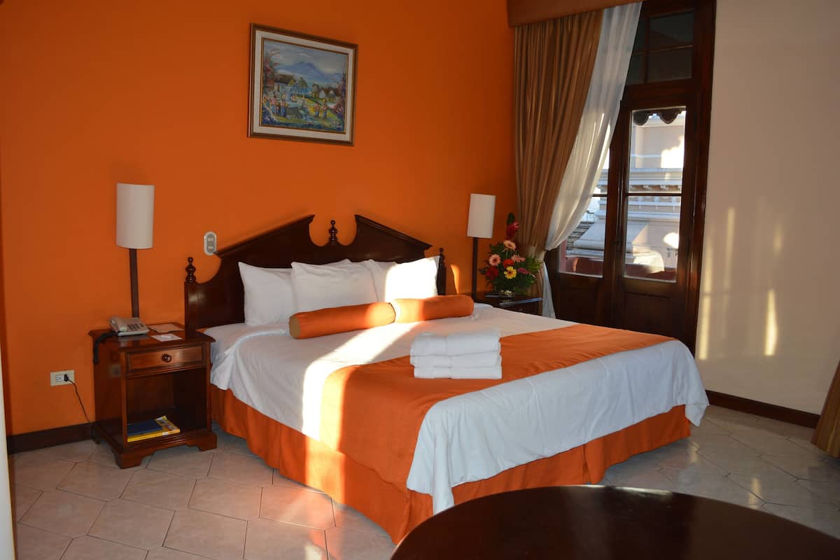 Une chambre d’hôtel avec un grand lit, une table de chevet avec une lampe, un tableau au mur et une fenêtre avec des rideaux.