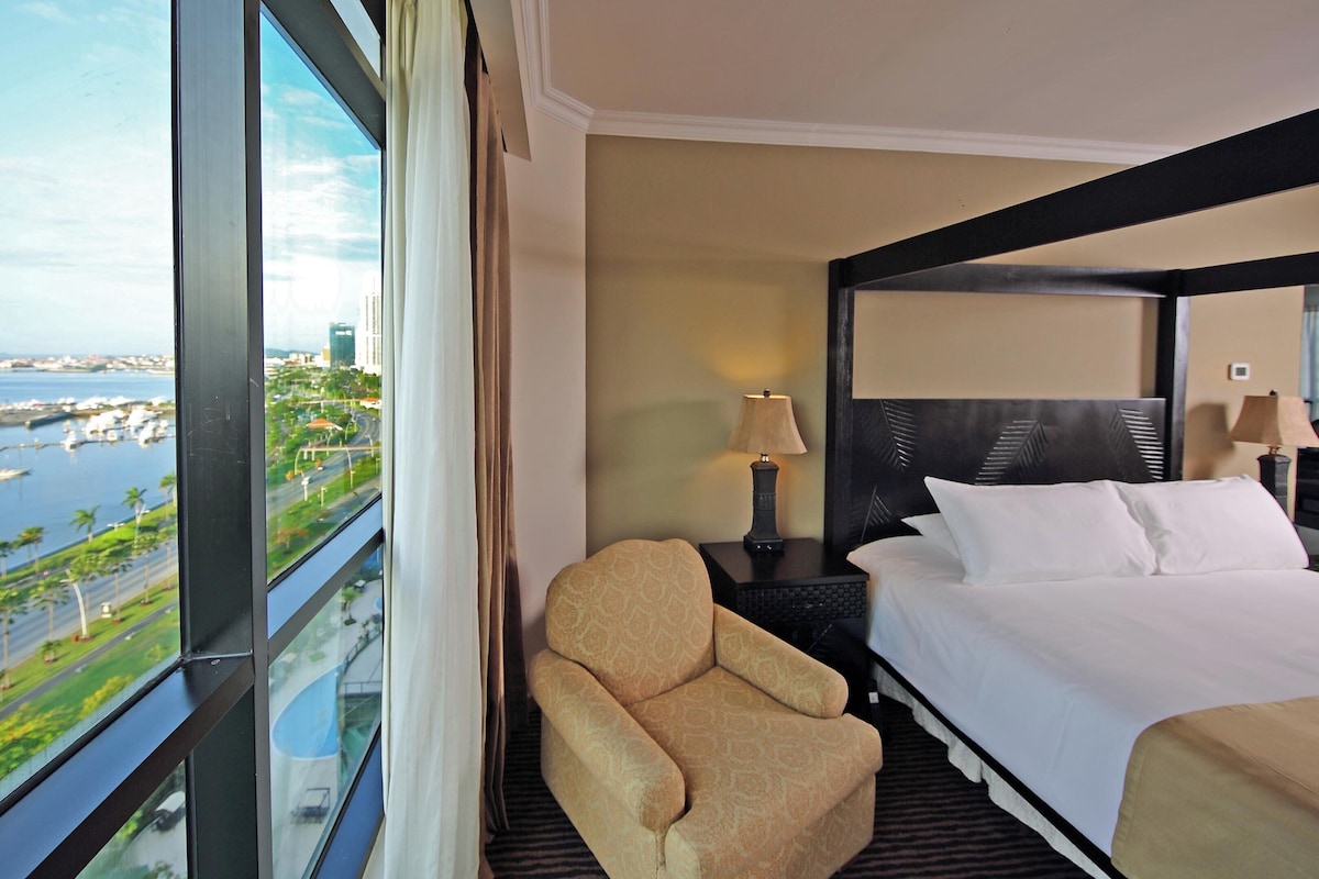 Una habitación de hotel con un ventanal grande, una cama, una silla y una mesita de noche con lámpara.
