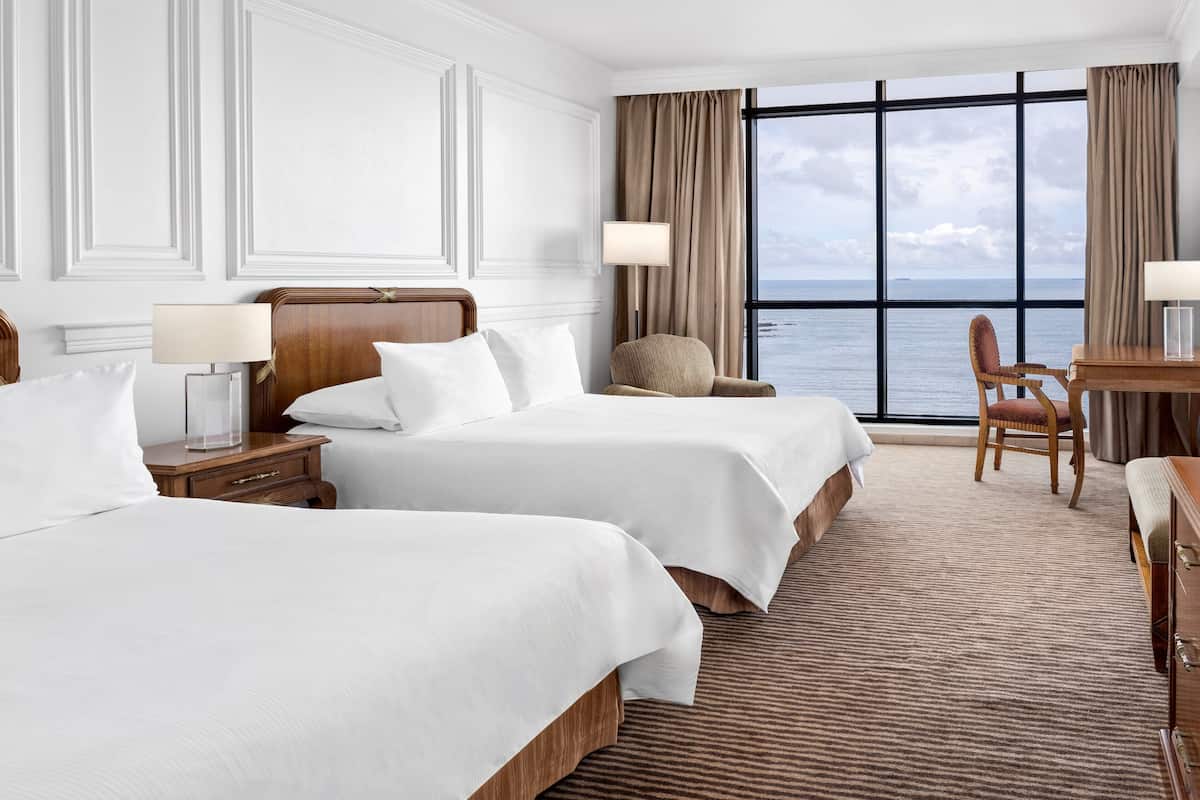 A hotel room with two beds, a desk, a chair, and a large window with a view of the sea.