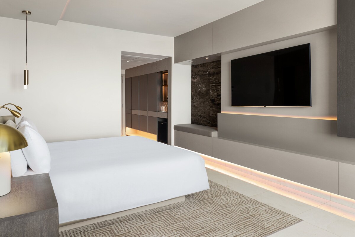 A modern bedroom with a large bed, a flat-screen TV mounted on the wall, and a built-in bench with a marble-like texture.