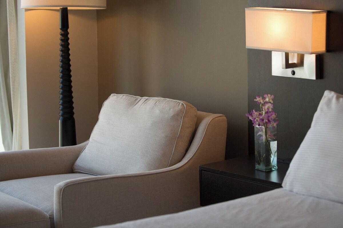 A hotel room with a bed, a chair, a nightstand with a lamp, and a vase with flowers.