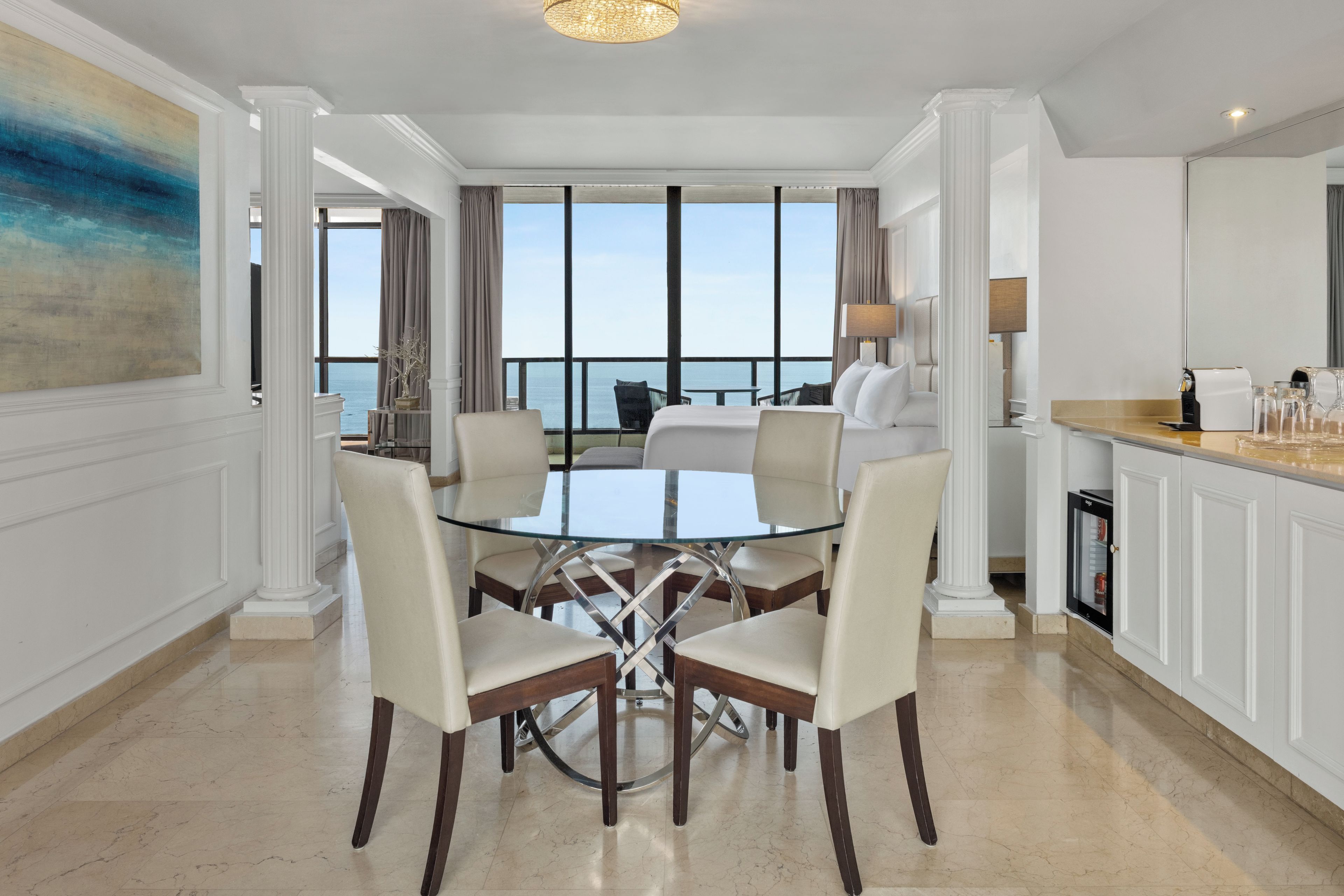Habitación de hotel con zona de comedor que incluye una mesa de vidrio y sillas, cama con ropa blanca, vista al mar y un minibar.