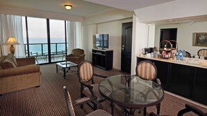 Habitación de hotel con balcón, sofá, mesa de comedor, sillas, televisión, minibar y vista al mar.