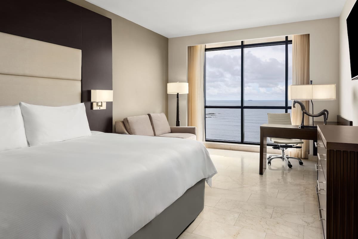 A hotel room with a large bed, a desk, a chair, and a view of the sea through a large window.