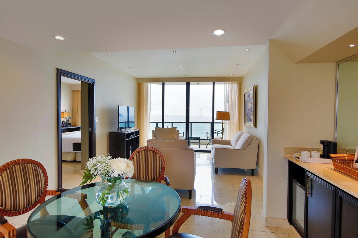 A hotel room with a dining area, a living room with a sofa and armchairs, a kitchen area, and a large window with a view.