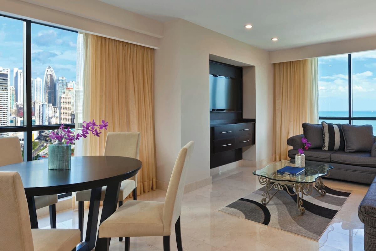 A modern living room with a dining area, a sofa set, a TV, and a view of the cityscape.
