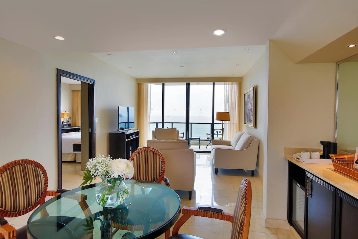 A hotel room with a dining area, a living room with a sofa and armchairs, a kitchen area, and a large window with a view.