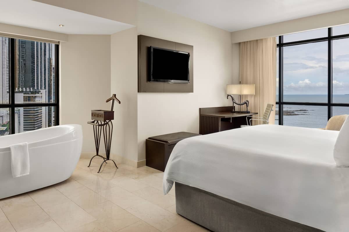 A hotel room with a large bed, a bathtub, a television mounted on the wall, a desk with a lamp, and a view of the ocean.